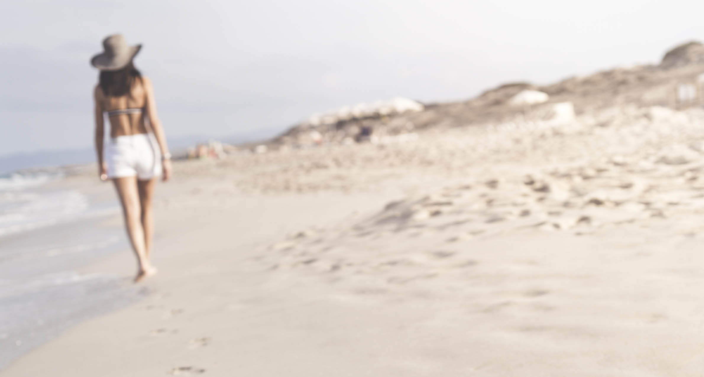 Distant Woman in shorts walking barefoot on white sandy beach in Formentera, Ibiza leaving footprints in sand. Focus is on the foreground footprints