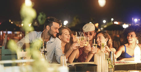 Happy friends cheering and drinking cocktails at beach party outdoor - Young millennials people having fun at weekend summer night - Youth lifestyle and nightlife concept - Main focus on left guys