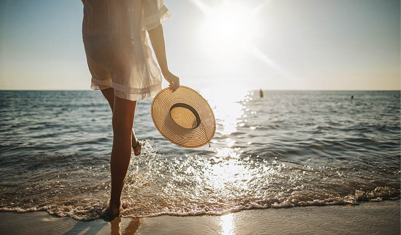Close-up of young woman in white sun dress and with hat in hand walking alone on sandy beach at summer sunset, splashing water in sea shallow
