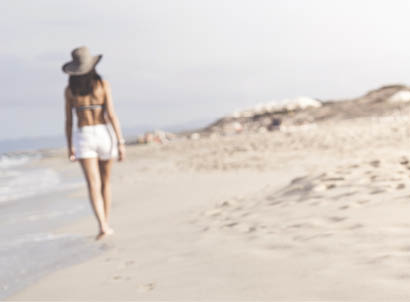 Distant Woman in shorts walking barefoot on white sandy beach in Formentera, Ibiza leaving footprints in sand. Focus is on the foreground footprints