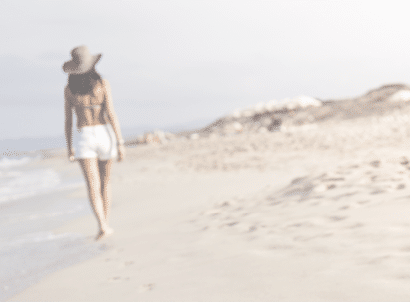 Distant Woman in shorts walking barefoot on white sandy beach in Formentera, Ibiza leaving footprints in sand. Focus is on the foreground footprints