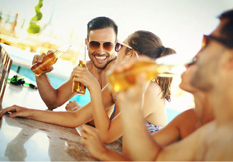 Group of mid 20's people having fun at beach bar. There are two guys and two girls on vacation together. One of the guys is in focus,facing camera and smiling. Each person is having beer and wearing sunglasses.