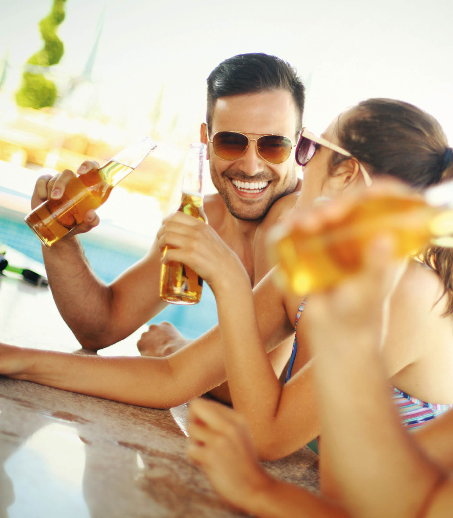 Group of mid 20's people having fun at beach bar. There are two guys and two girls on vacation together. One of the guys is in focus,facing camera and smiling. Each person is having beer and wearing sunglasses.
