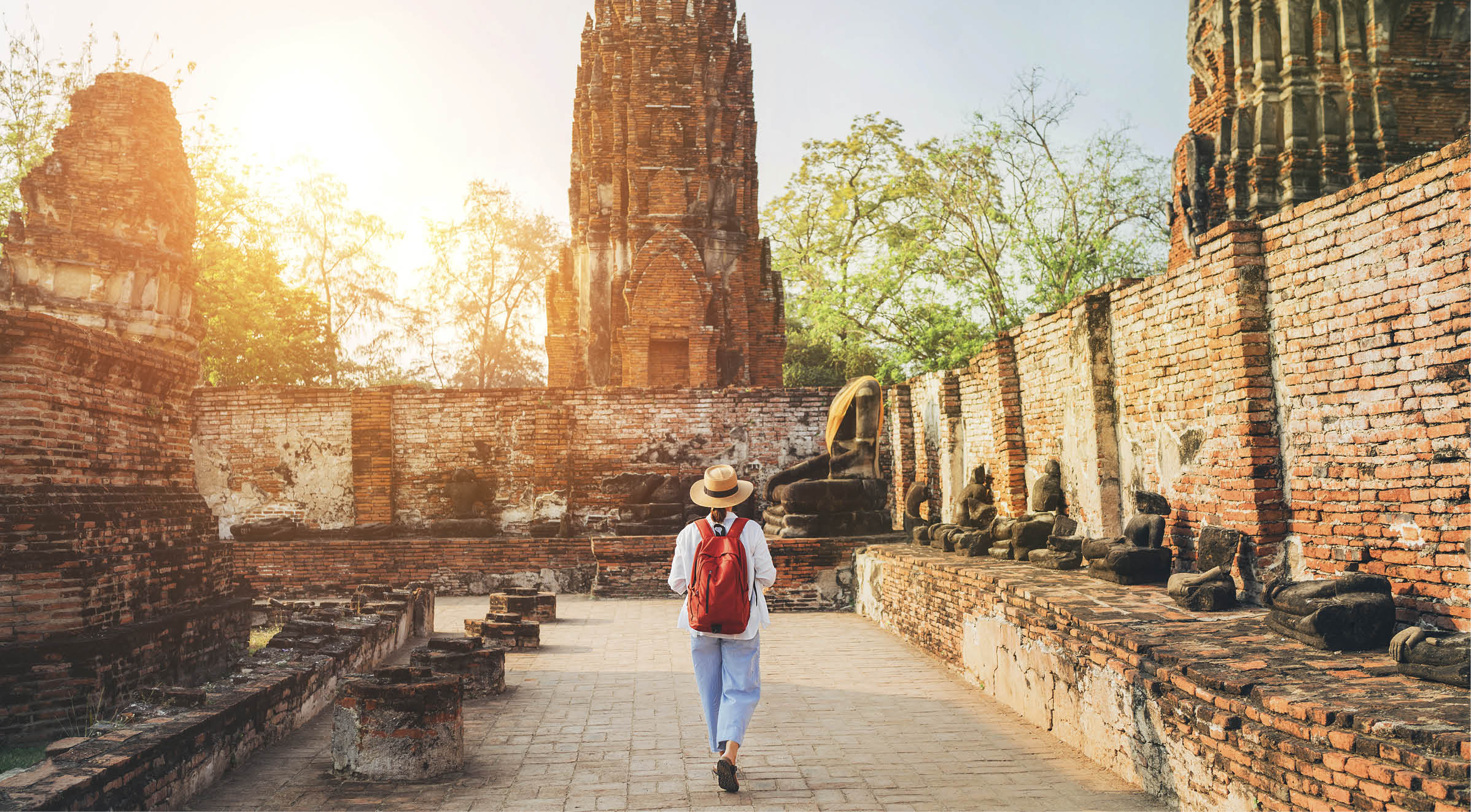 Young woman tourist in a straw hat and white clothes walking with light city backpack through Ayutthaya Wat Phra Ram ancient ruins streets in Thailand. History, tourism, sightseeing concept.