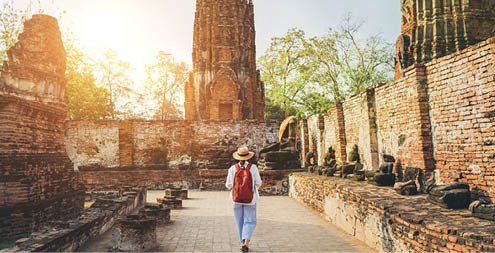 Young woman tourist in a straw hat and white clothes walking with light city backpack through Ayutthaya Wat Phra Ram ancient ruins streets in Thailand. History, tourism, sightseeing concept.