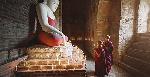 Two young Burmese buddhist monks in their typical clothing inside stupa temple praying and worshipping buddha. Natural Light inside dark temple with giant Buddha Statue. Bagan, Mandalay Region, Myanmar.