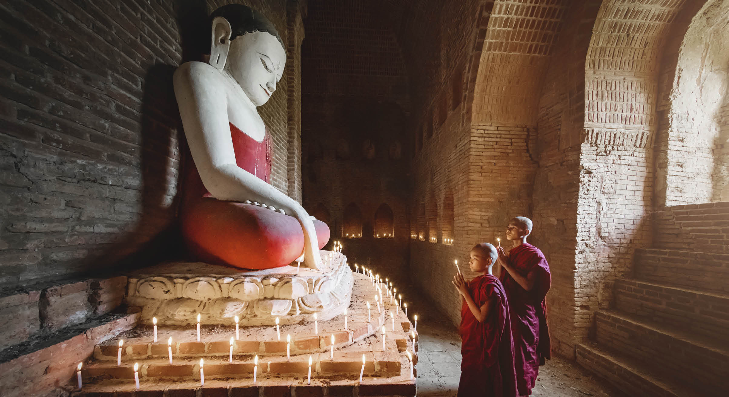 Two young Burmese buddhist monks in their typical clothing inside stupa temple praying and worshipping buddha. Natural Light inside dark temple with giant Buddha Statue. Bagan, Mandalay Region, Myanmar.