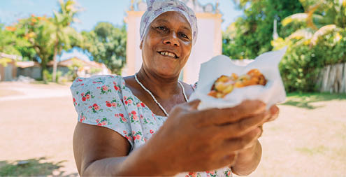 Happy Brazilian woman dressed in the traditional Bahian costume of the Umbanda religion, offering acarajÃ  - typical Bahia food - in the historic center of Porto Seguro in the background