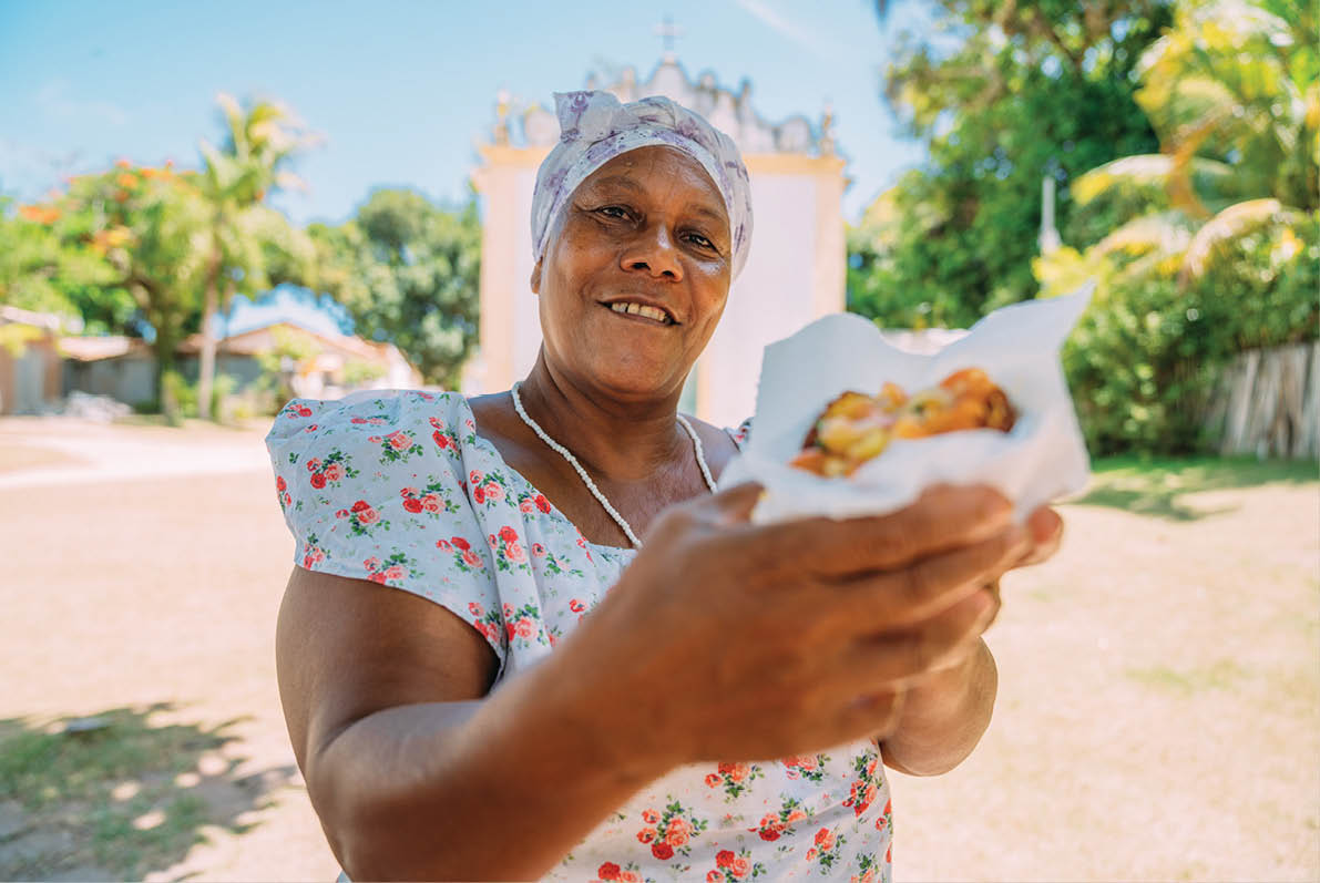 Happy Brazilian woman dressed in the traditional Bahian costume of the Umbanda religion, offering acarajÃ  - typical Bahia food - in the historic center of Porto Seguro in the background