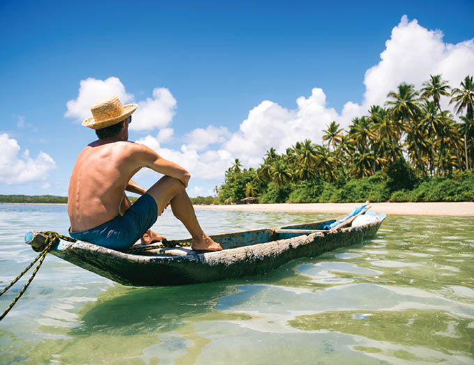 Tourist wearing a straw sun hat sitting in a rustic dugout canoe on a sunny palm-lined tropical island beach in Bahia, Brazil